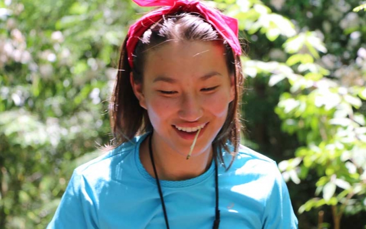 A young person smiles with a toothpick in their mouth. There are green leaves blurred behind them. 